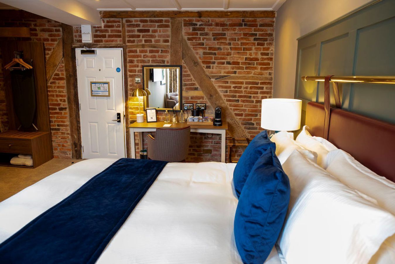A hotel room with a brick wall, a desk with a mirror, a bed with blue and white bedding, and a wooden headboard.
