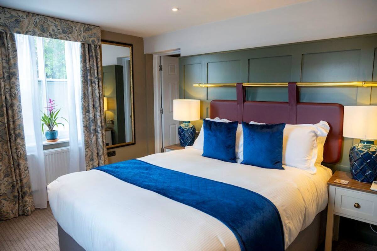 Una habitación de hotel con una cama grande, dos mesitas de noche, una lámpara de noche, una ventana con cortinas y una planta en maceta.