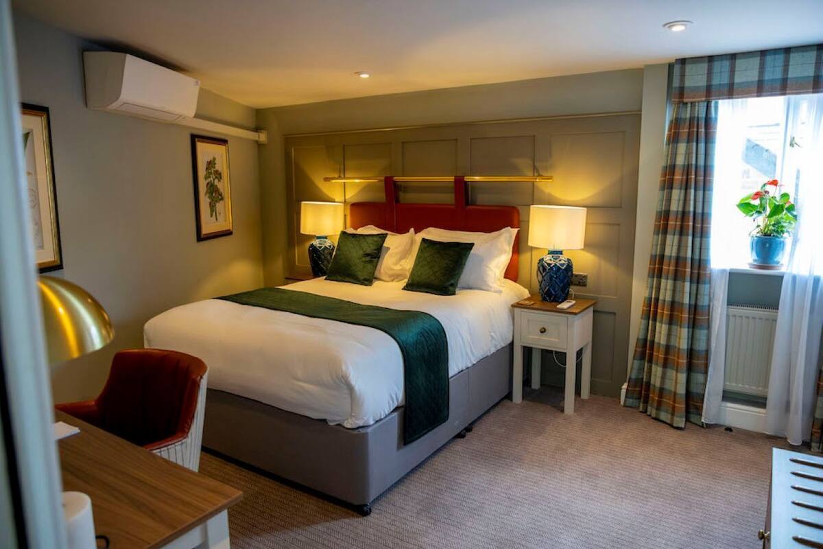 Habitación de hotel con cama, mesitas de noche, escritorio, silla, ventana con cortinas y radiador.
