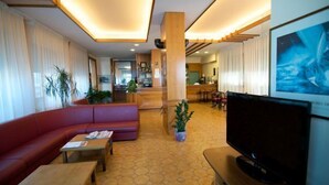 Un vestíbulo de hotel con un sofá rojo, una mesa de centro de madera y un televisor. Hay plantas y un suelo de baldosas.