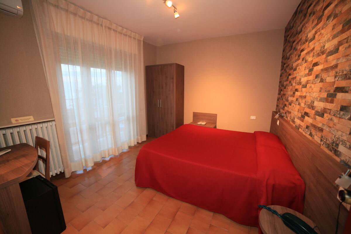 A bedroom with a red bedspread, a wooden wardrobe, a desk with a chair, and a radiator.