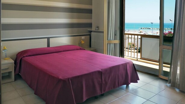 Habitación de hotel con una cama, una mesita de noche, una lámpara y vistas a la playa desde un balcón.