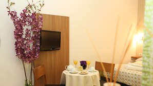 Habitación de hotel con cama, televisor montado en la pared, mesa de comedor con artículos de desayuno y una planta de orquídea en maceta.