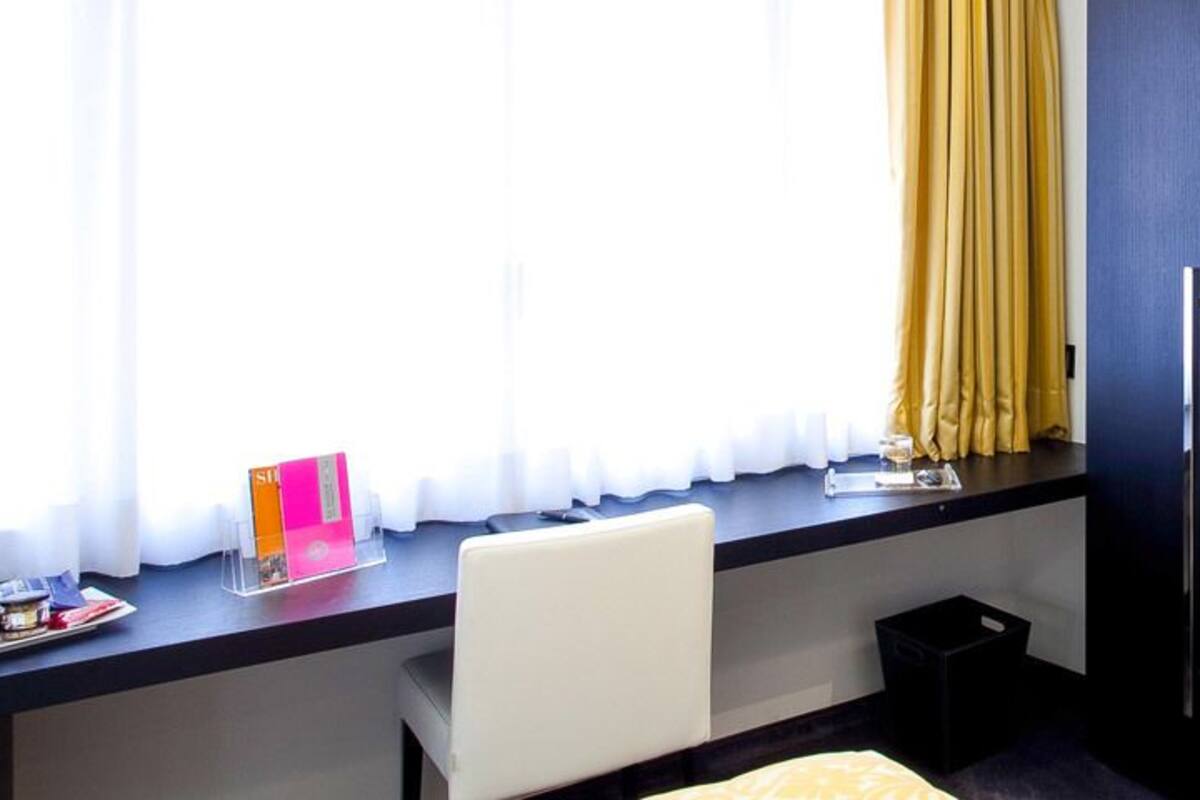Una habitación de hotel moderna con escritorio, dos sillas, una cama con almohada amarilla y un ventanal con cortinas.