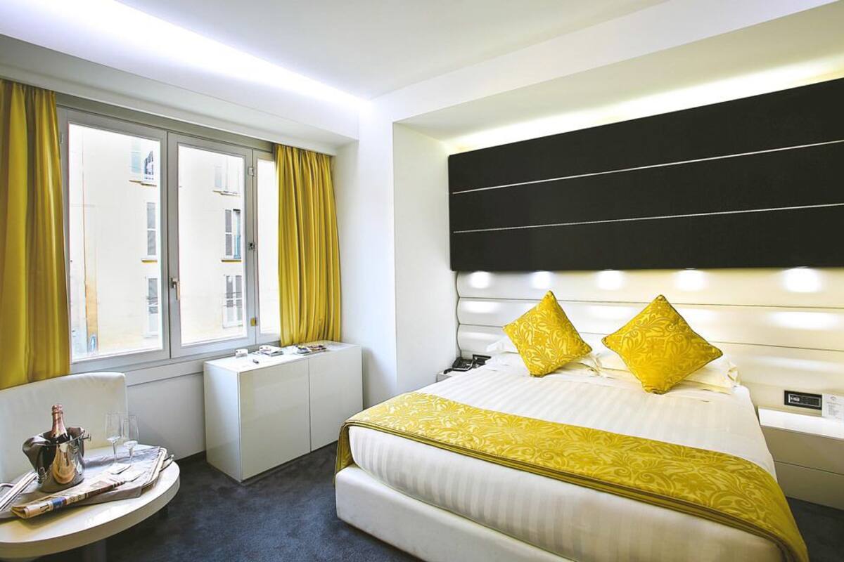Habitación de hotel con una cama grande, una mesita de noche, una silla, una ventana con cortinas y vistas a edificios.