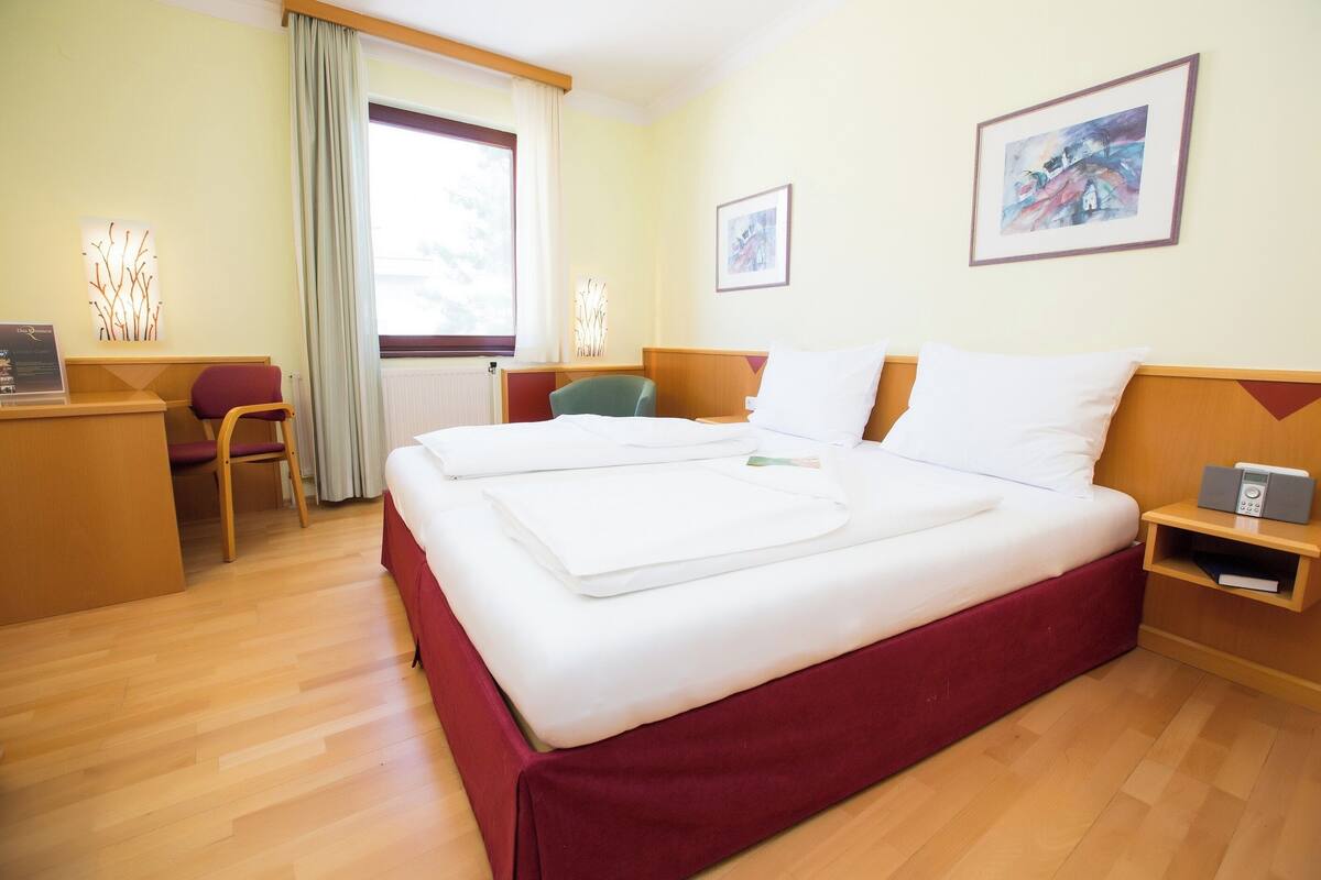 A hotel room with a large bed, a desk, a chair, and two framed pictures on the walls.