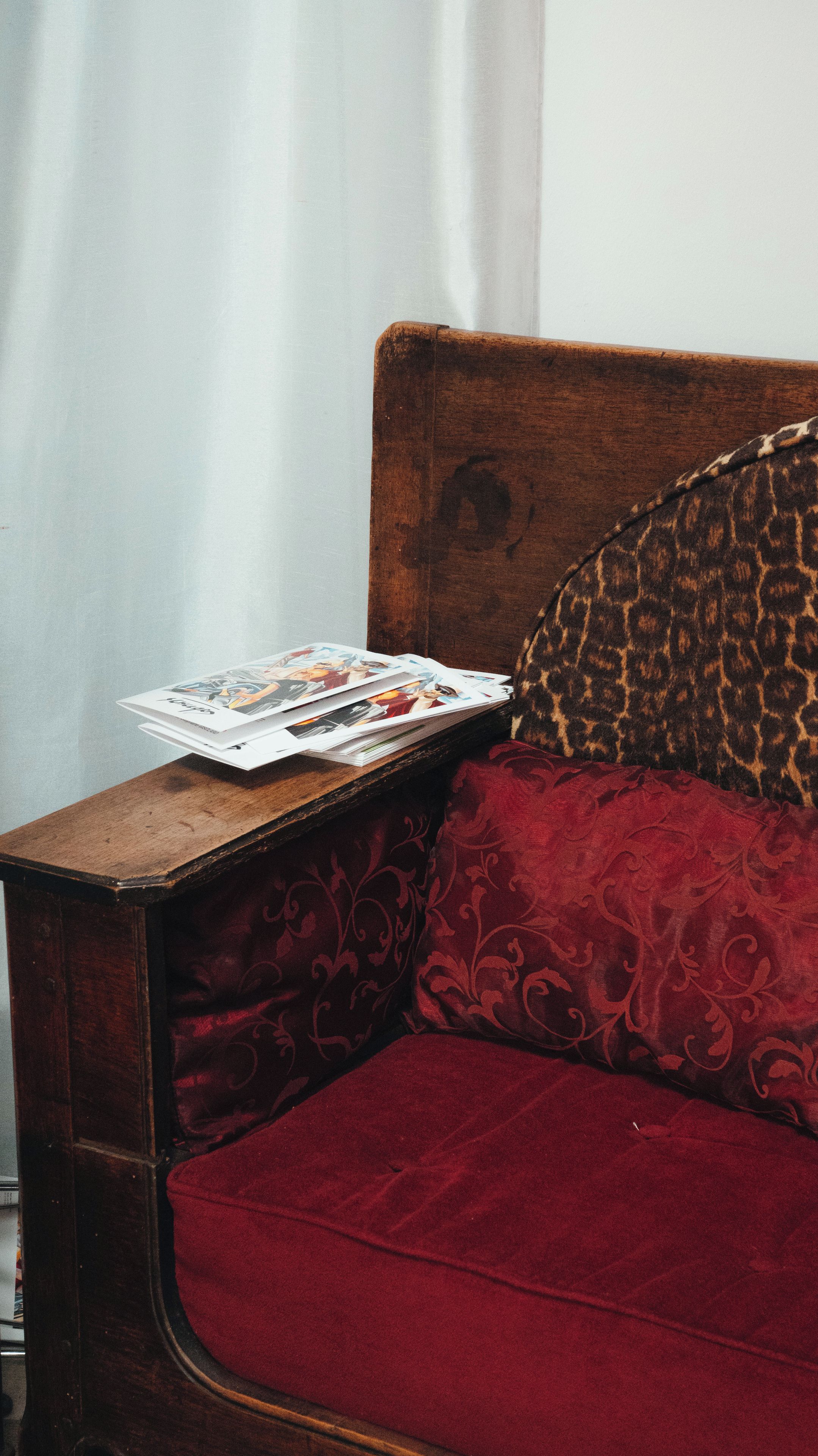 Cabecera de madera con almohada tapizada en rojo y una pila de revistas sobre una mesita de noche de madera.