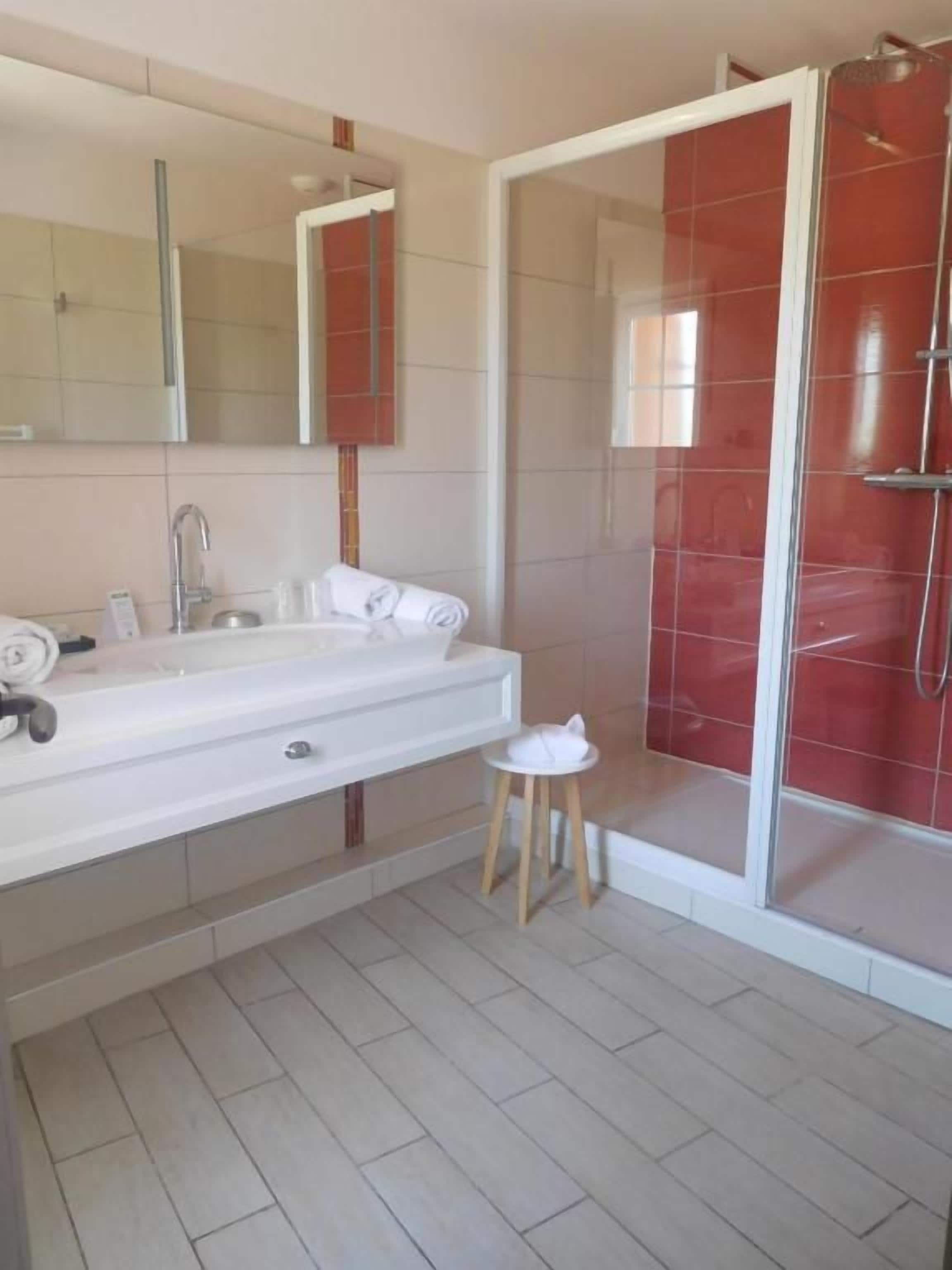 A bathroom with a white sink, a mirror, a shower with red tiles, and a small stool.