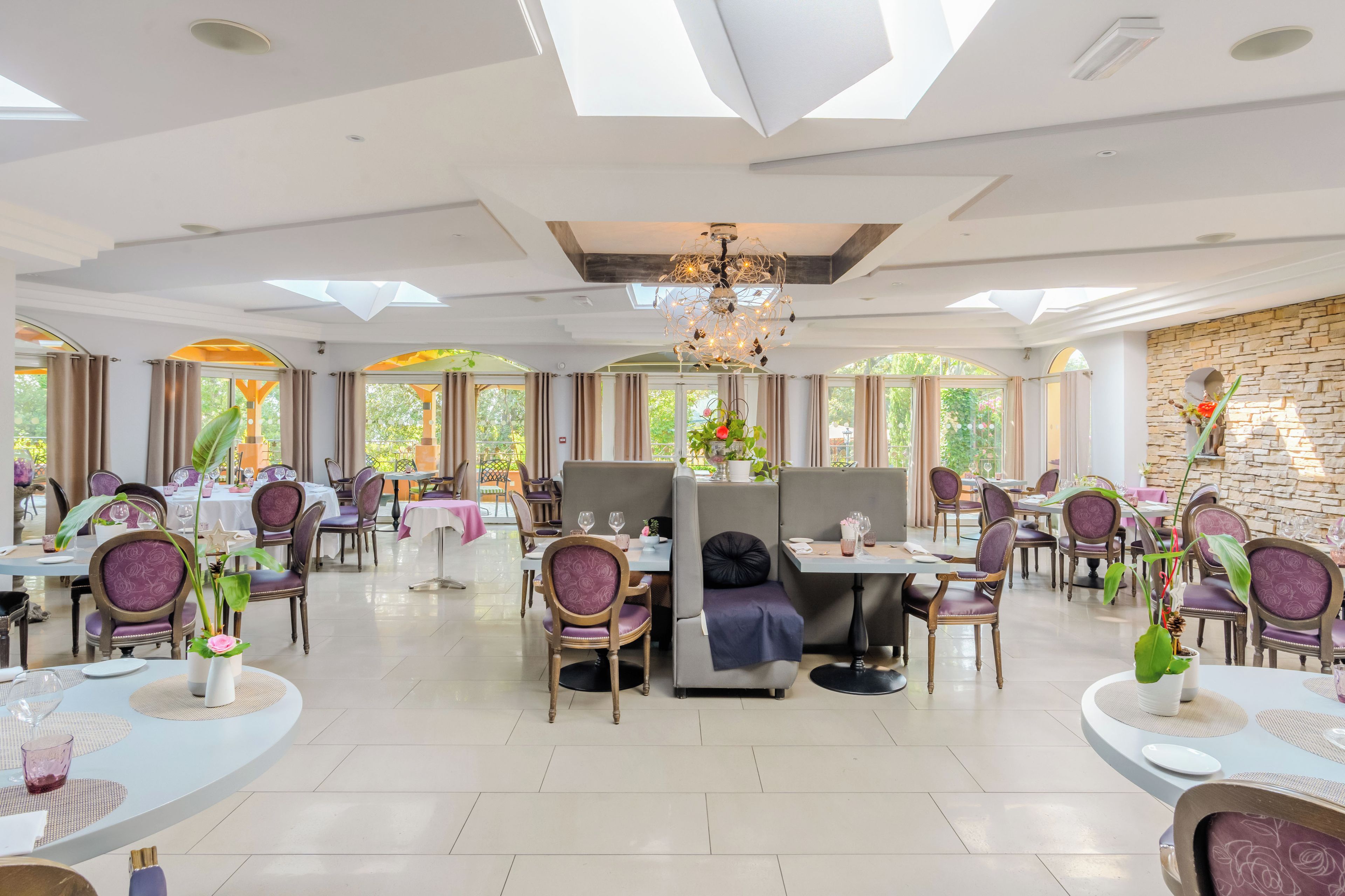 Un vaste espace dédié à la salle à manger, avec de nombreuses tables et chaises, un lustre et de grandes fenêtres laissant entrer la lumière naturelle.
