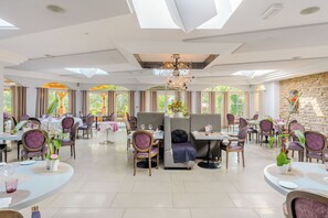 Un'ampia sala da pranzo con diversi tavoli e sedie, un lampadario e ampie finestre che lasciano entrare la luce naturale.