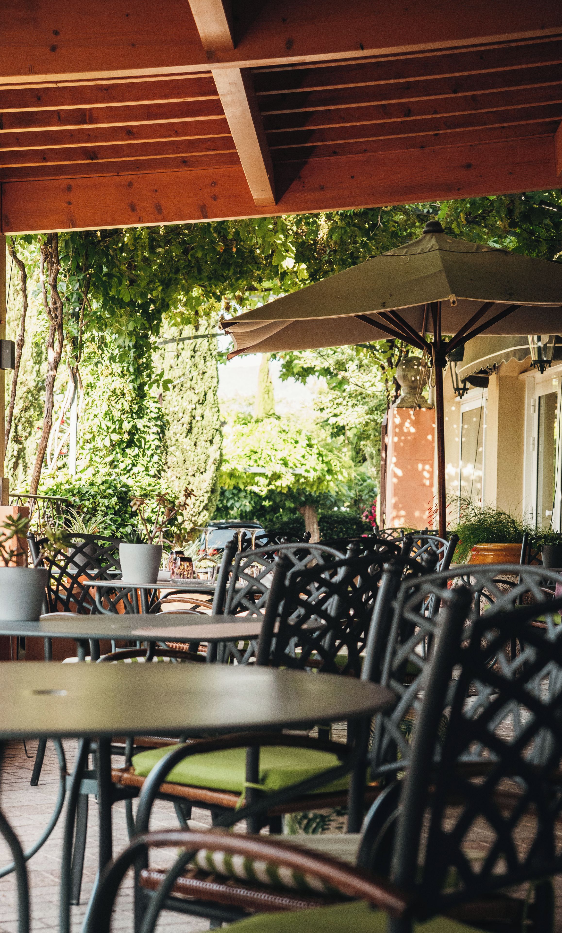 An outdoor cafe with tables and chairs, surrounded by greenery and a wooden structure overhead.