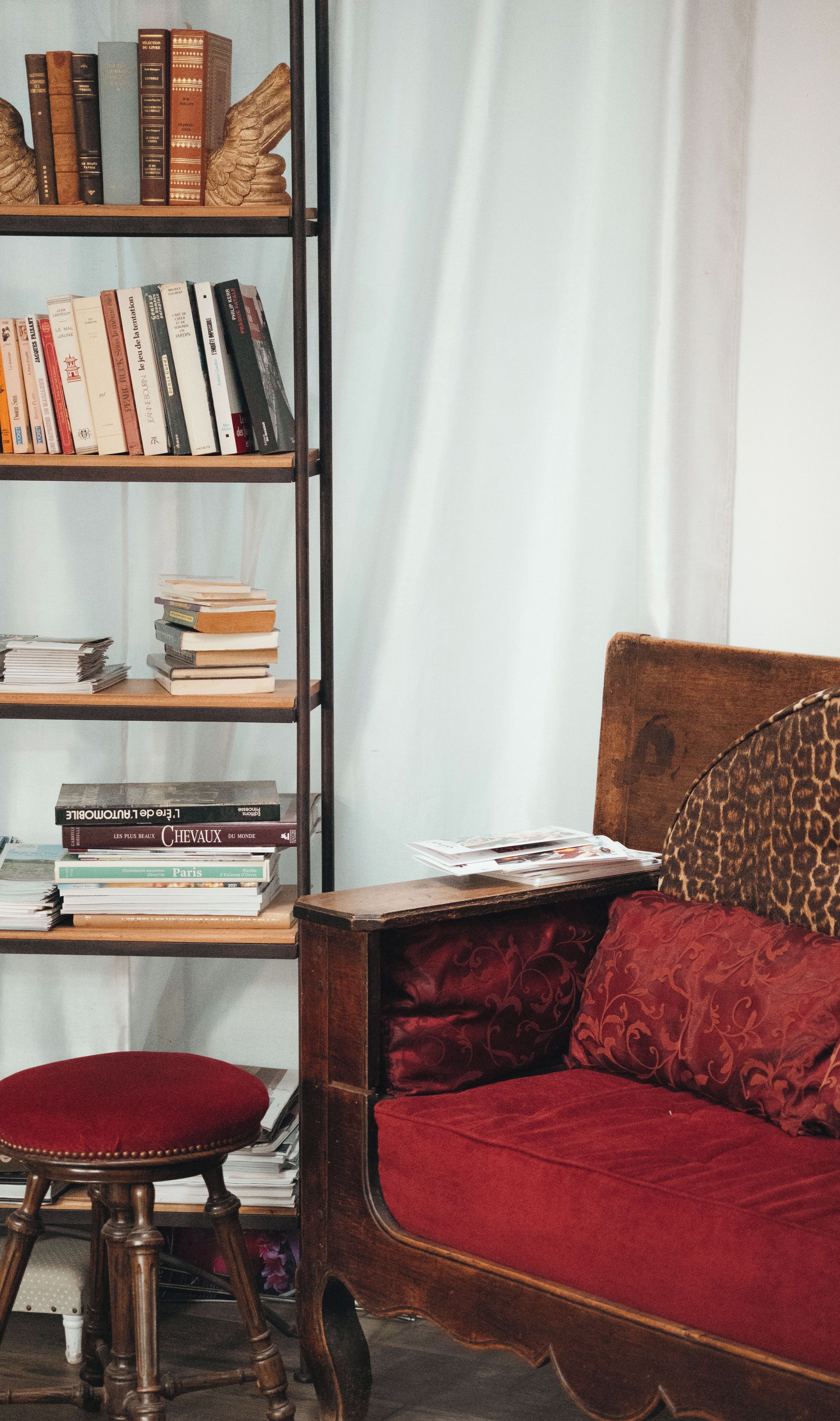 Ein Zimmer mit einem Holzregal voller Bücher, einem roten Samtsofa und einem passenden Hocker.