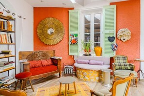 A room with a sofa, a small table, a bookshelf, and a window with shutters.