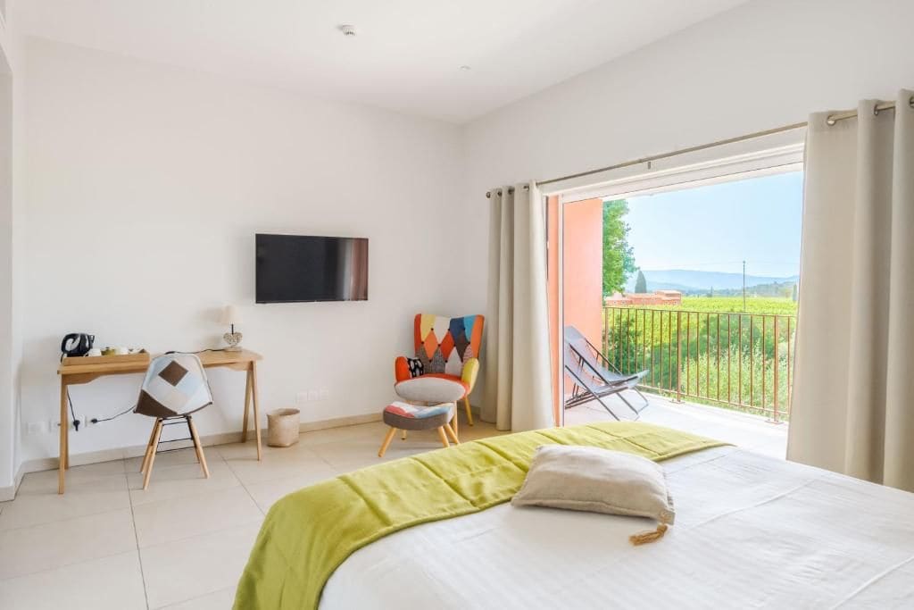 A hotel room with a bed, a desk with a chair, a television, and a balcony with a view of greenery.