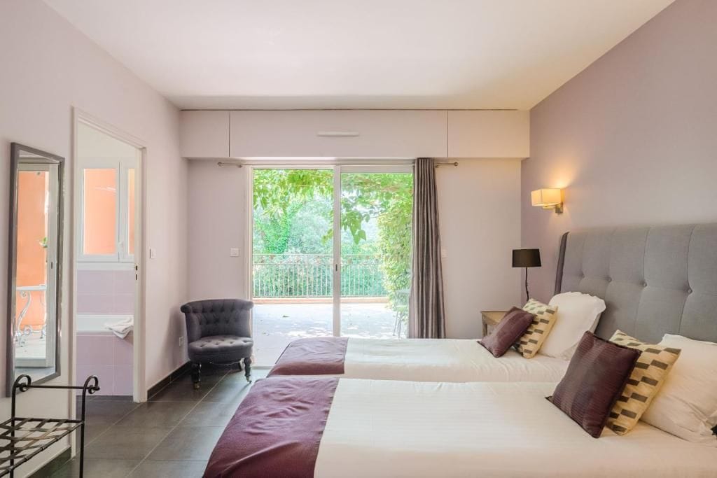 Une chambre d’hôtel avec deux lits, une chaise, une fenêtre donnant sur un espace verdoyant et une porte.