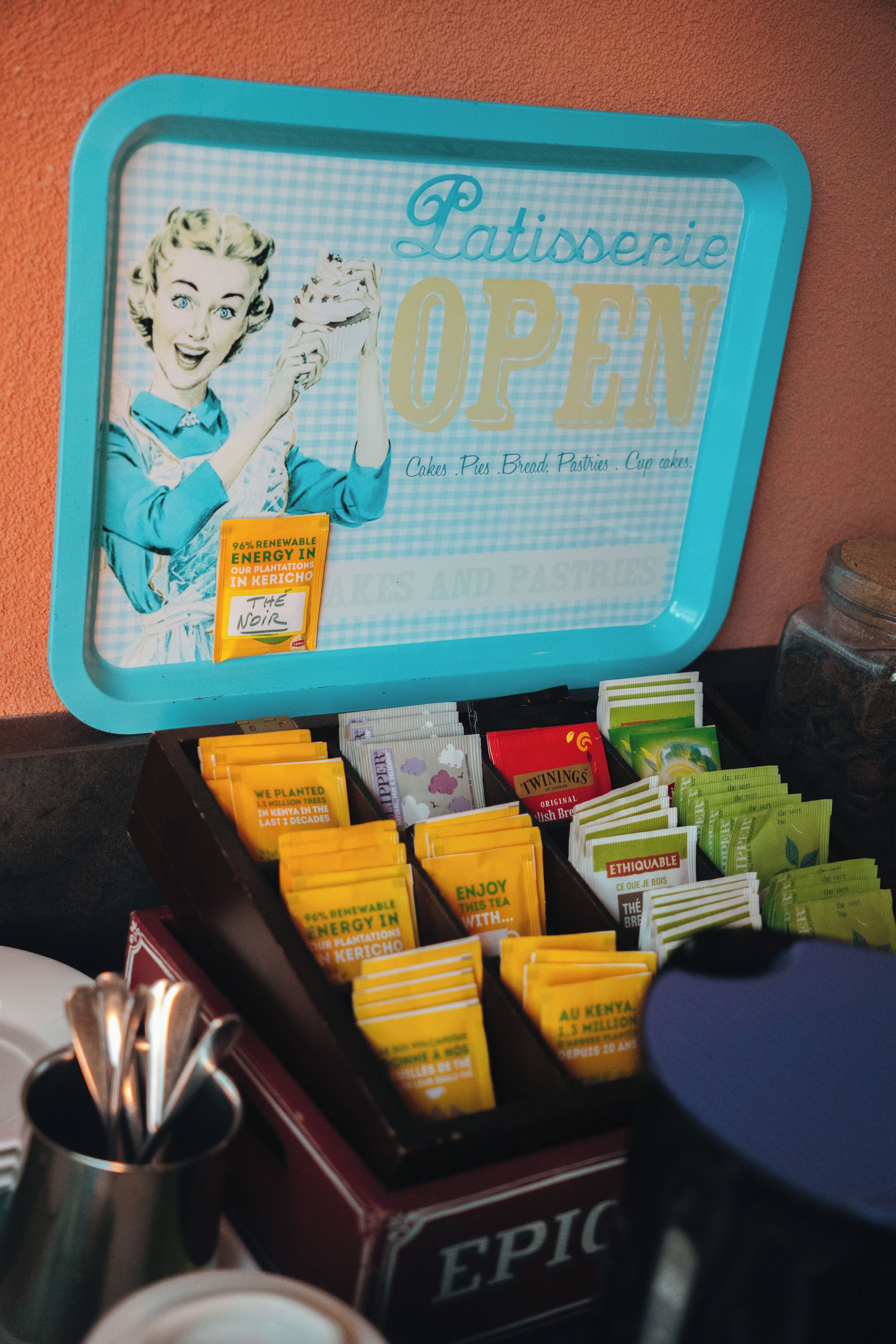 A tray with a vintage design, containing various tea bags and a sign indicating a patisserie is open.