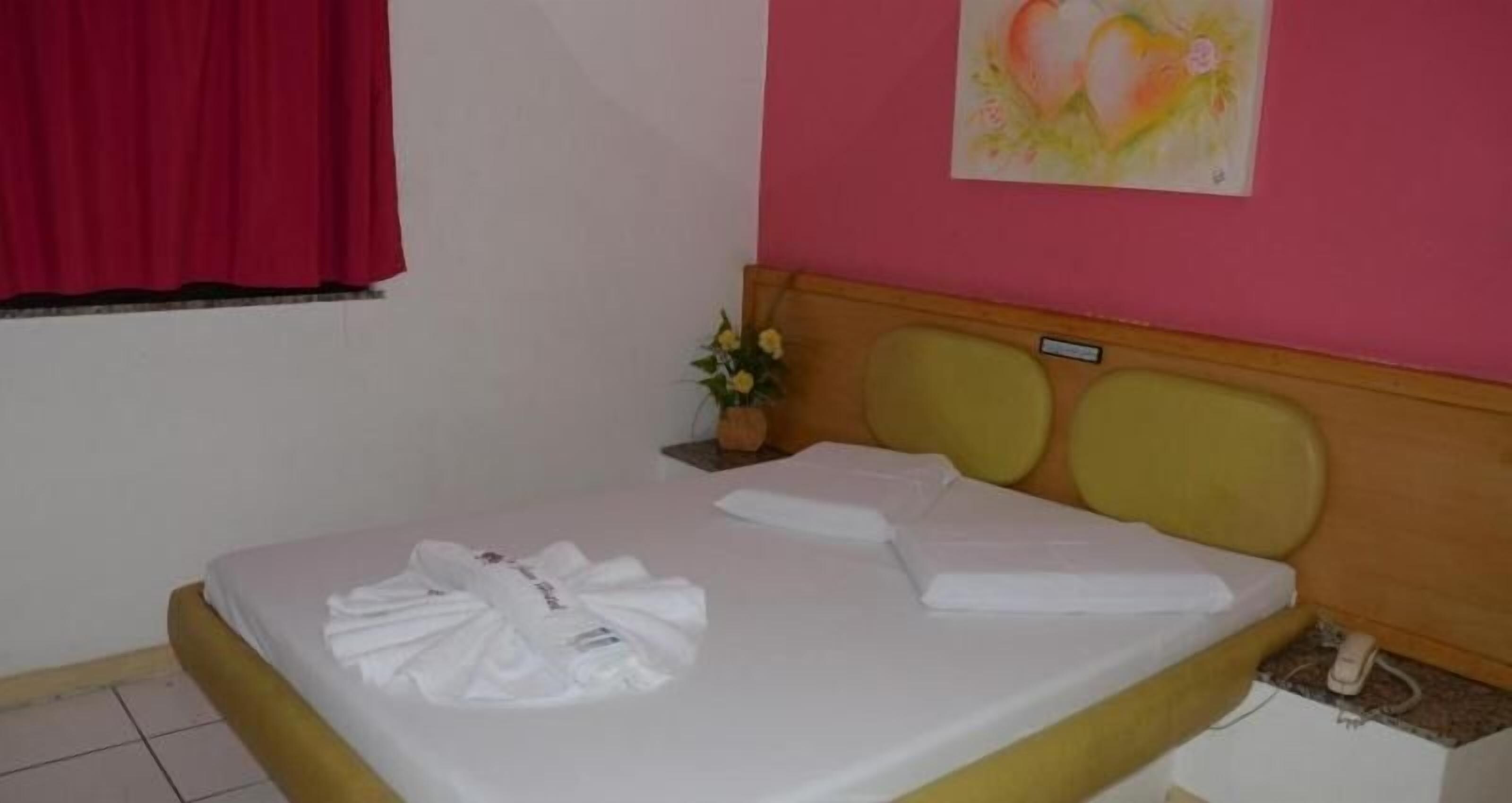 A neatly made bed with white linens, a small bedside table with a vase of flowers, and a painting of a heart on the wall.