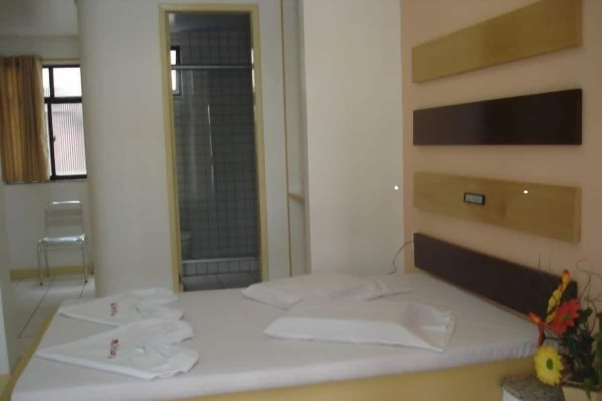 Ein Hotelzimmer mit Bett, Dusche, Stuhl und einer an der Wand befestigten Bedieneinheit.