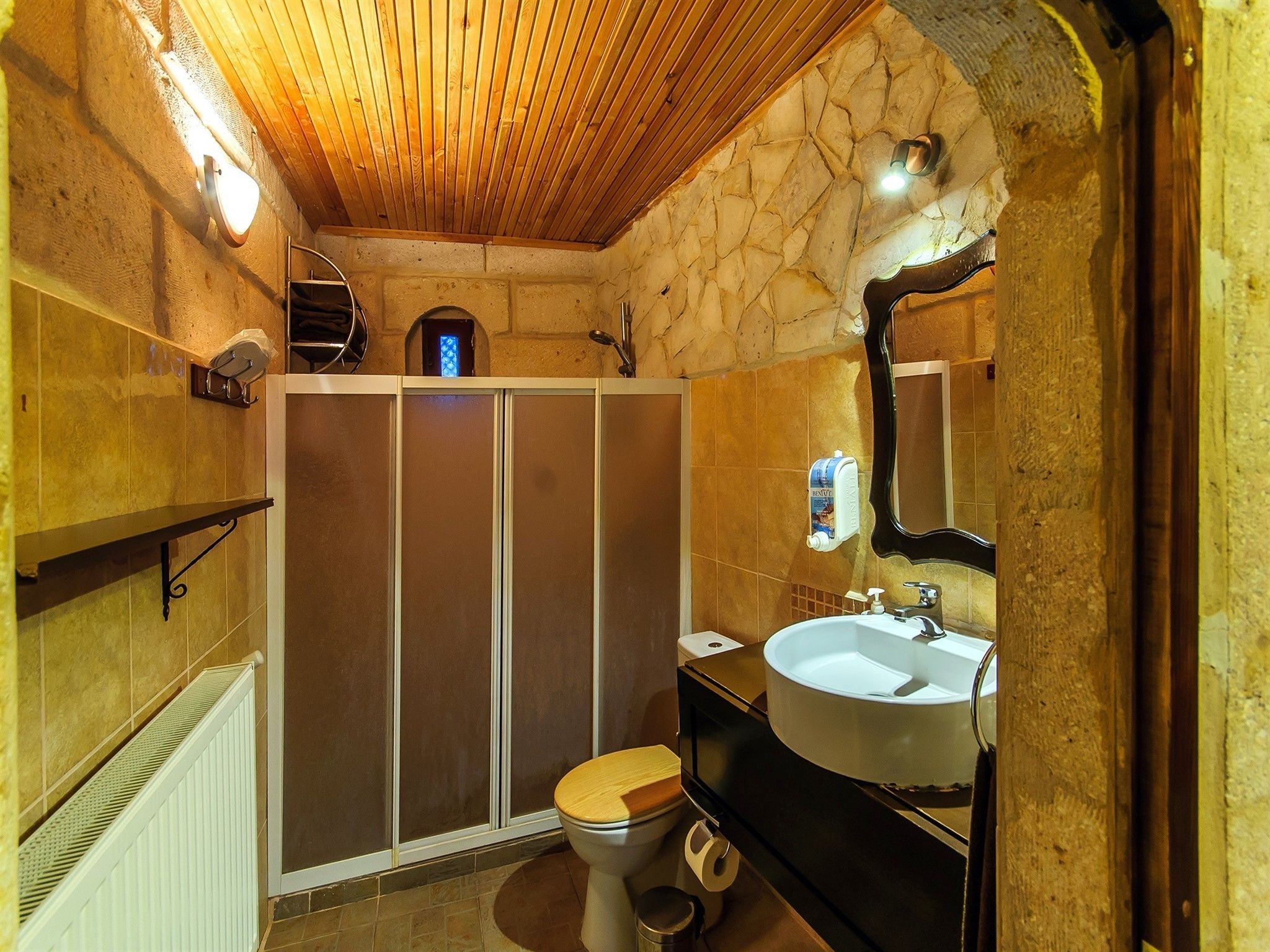 A bathroom with a wooden ceiling, stone walls, a wooden toilet, a sink with a mirror, and a shower enclosure.