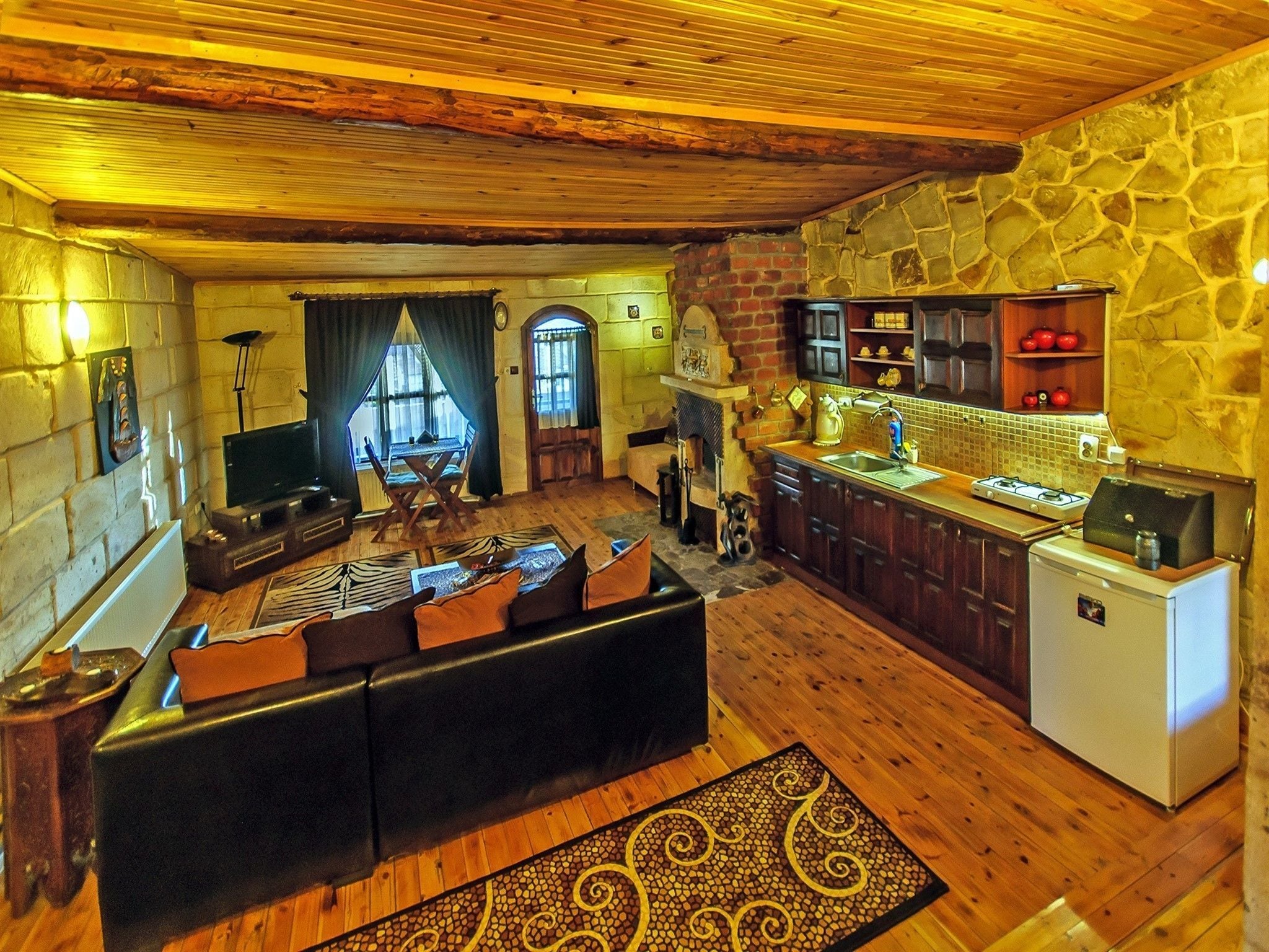 Una sala de estar acogedora con sofá, televisor y chimenea. Cuenta con una cocina equipada con refrigerador, estufa y armarios. El ambiente se complementa con pisos de madera y paredes de piedra.