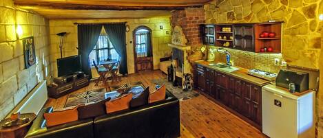 Una sala de estar acogedora con sofá, televisor y chimenea. Cuenta con una cocina equipada con refrigerador, estufa y armarios. El ambiente se complementa con pisos de madera y paredes de piedra.