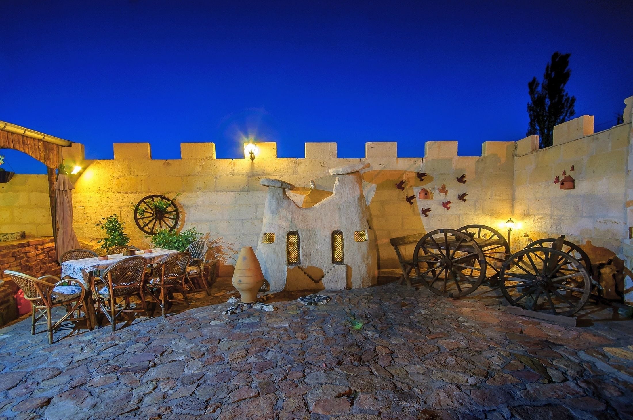 An outdoor dining area with stone flooring, rustic furniture, and a medieval-style wall with a wheel and small windows.