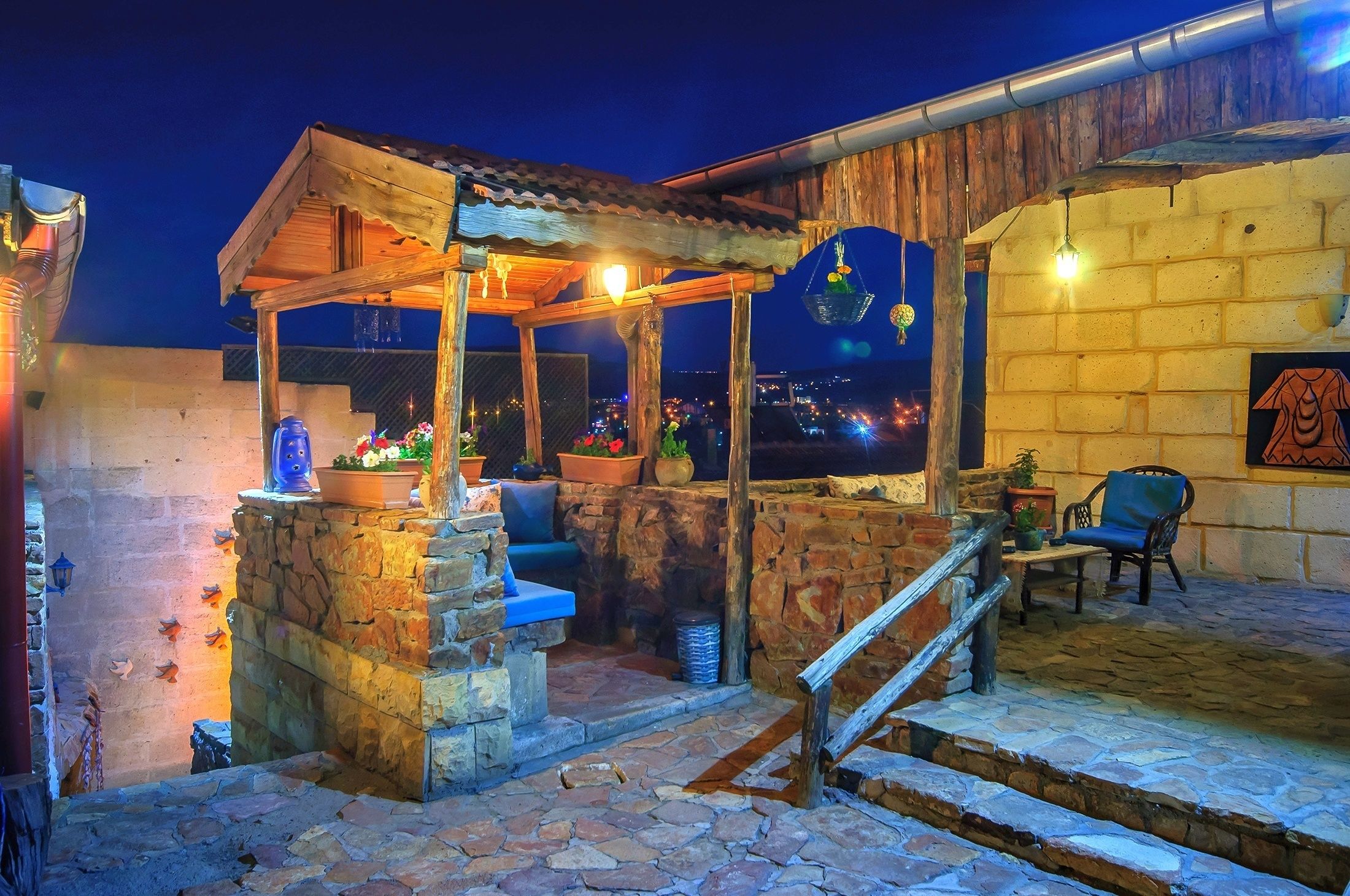 A rustic outdoor seating area with stone steps, a wooden pergola, and a view of the city at night.