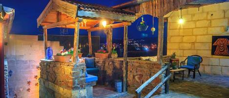 A rustic outdoor seating area with stone steps, a wooden pergola, and a view of the city at night.