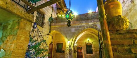Un patio con paredes de piedra, una mesa de madera y sillas. Se observan luces colgantes y una puerta al fondo.