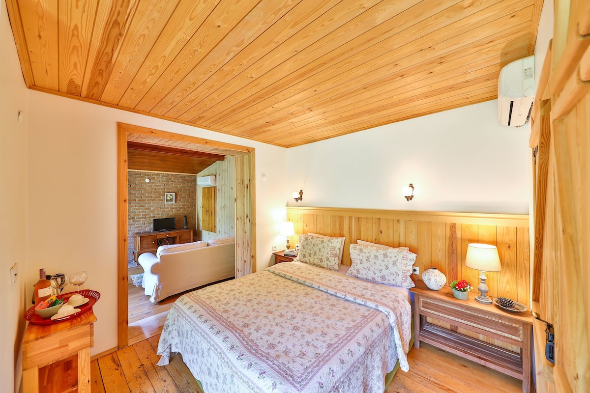 A cozy bedroom with a wooden ceiling, a bed with floral bedding, a nightstand with a lamp, and a small table with a bowl and glasses.