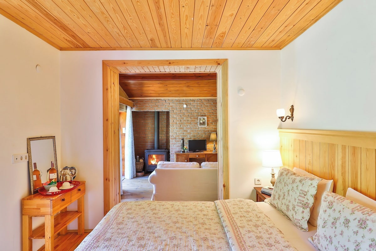 A cozy bedroom with a wooden ceiling, a brick fireplace, a bed with floral bedding, and a wooden nightstand with a lamp.