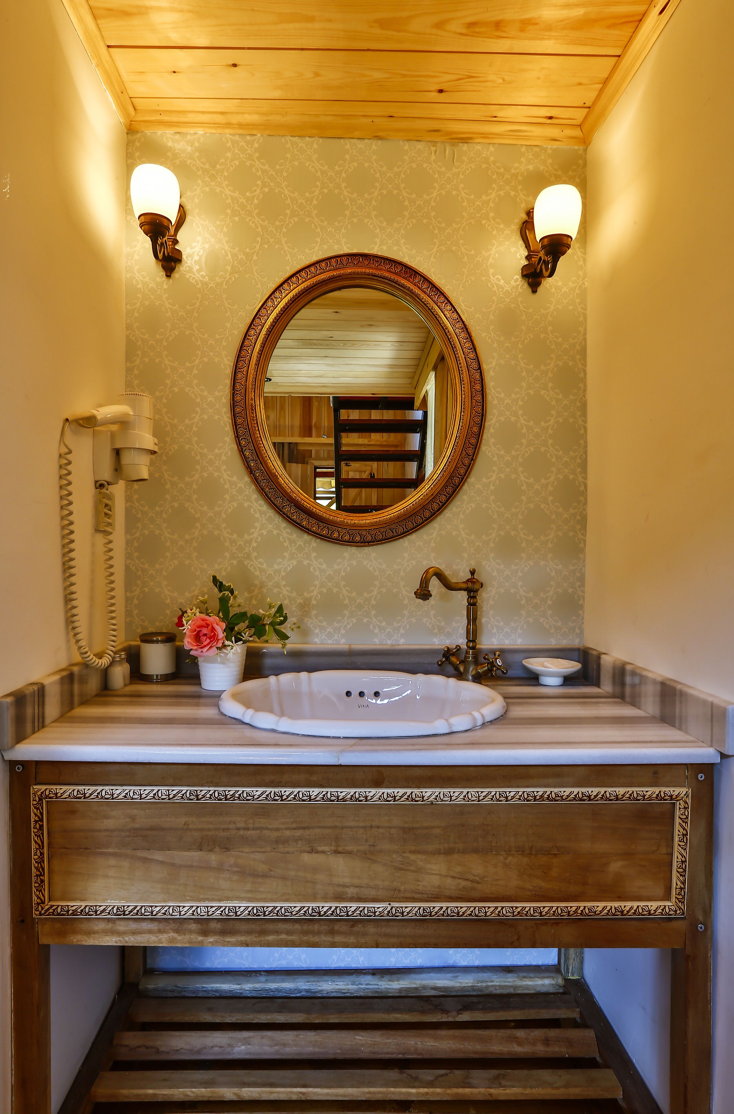 A bathroom with a wooden ceiling, a round mirror with a decorative frame, a white sink, a vintage-style faucet, and two wall-mounted light fixtures.