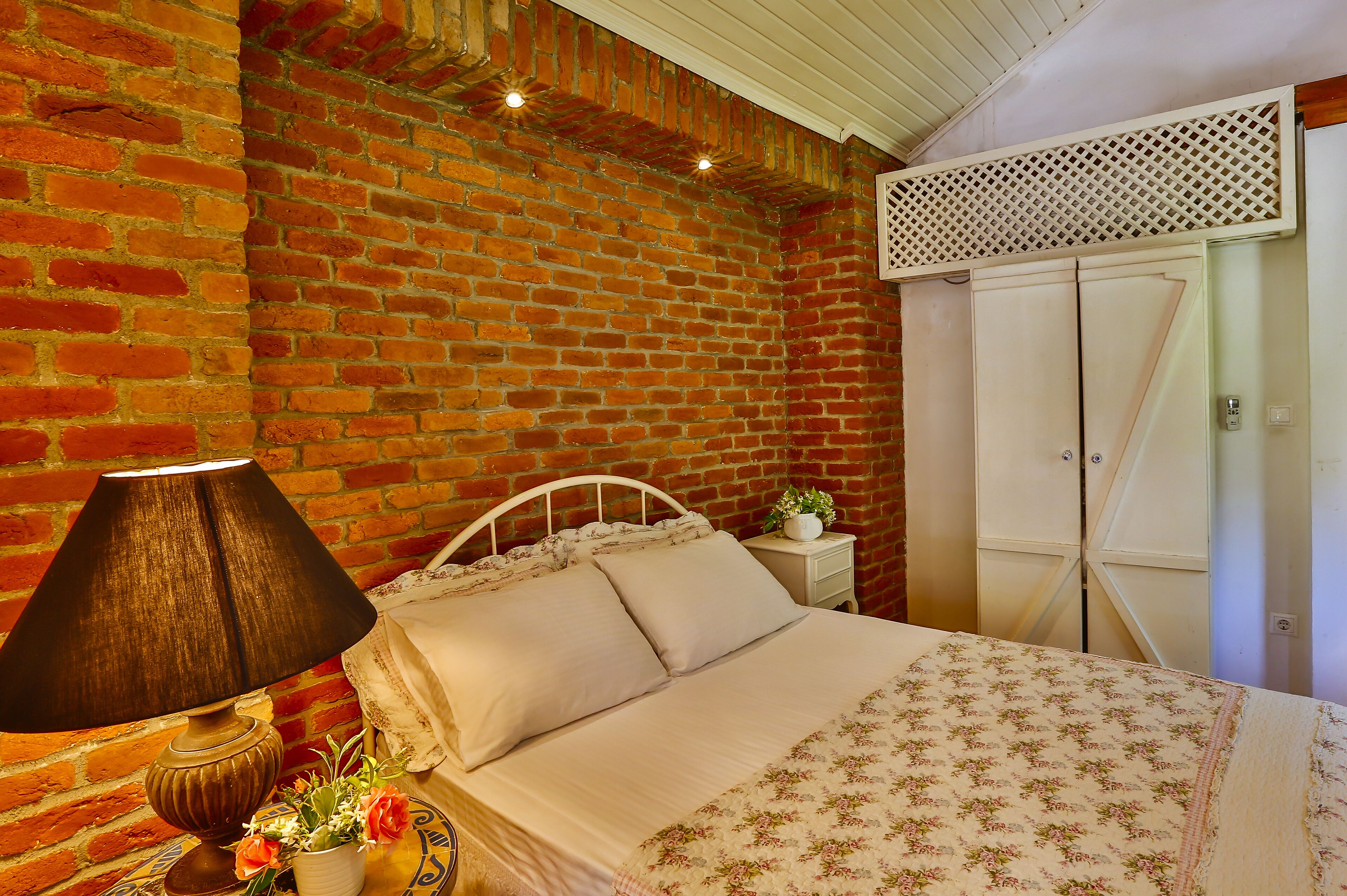 Un dormitorio con pared de ladrillo, una cama con colcha estampada, una lámpara y una mesita de noche con planta.