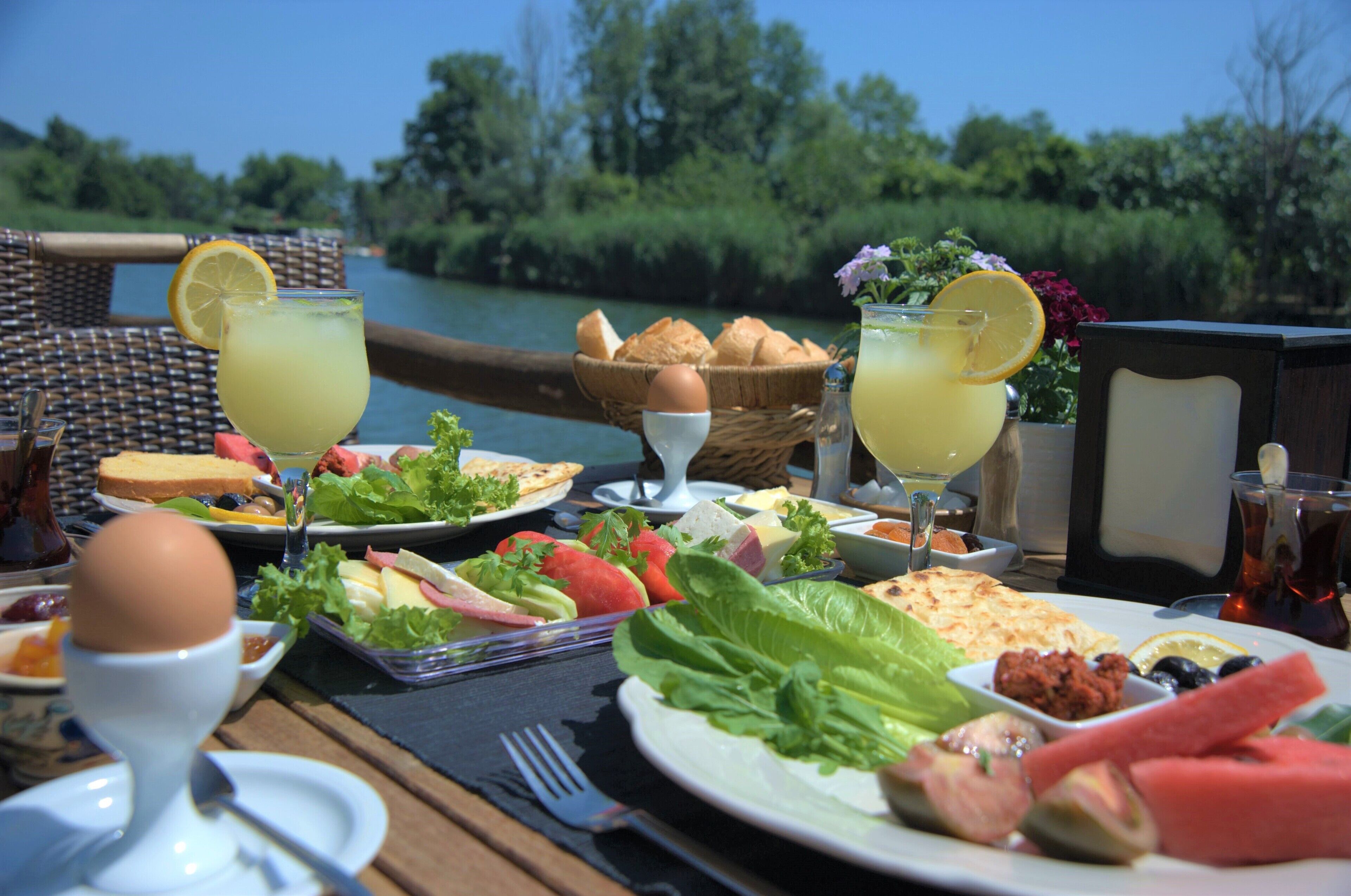 A table set with a variety of foods and drinks, including bread, eggs, and salads, with a view of a river and greenery in the background.