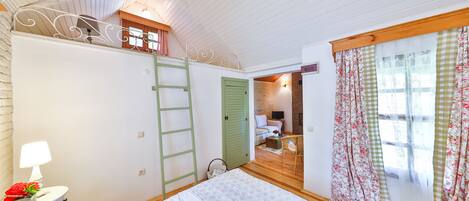 A bedroom with a bed, a dresser, a nightstand, a lamp, a window with floral curtains, and a door leading to another room.