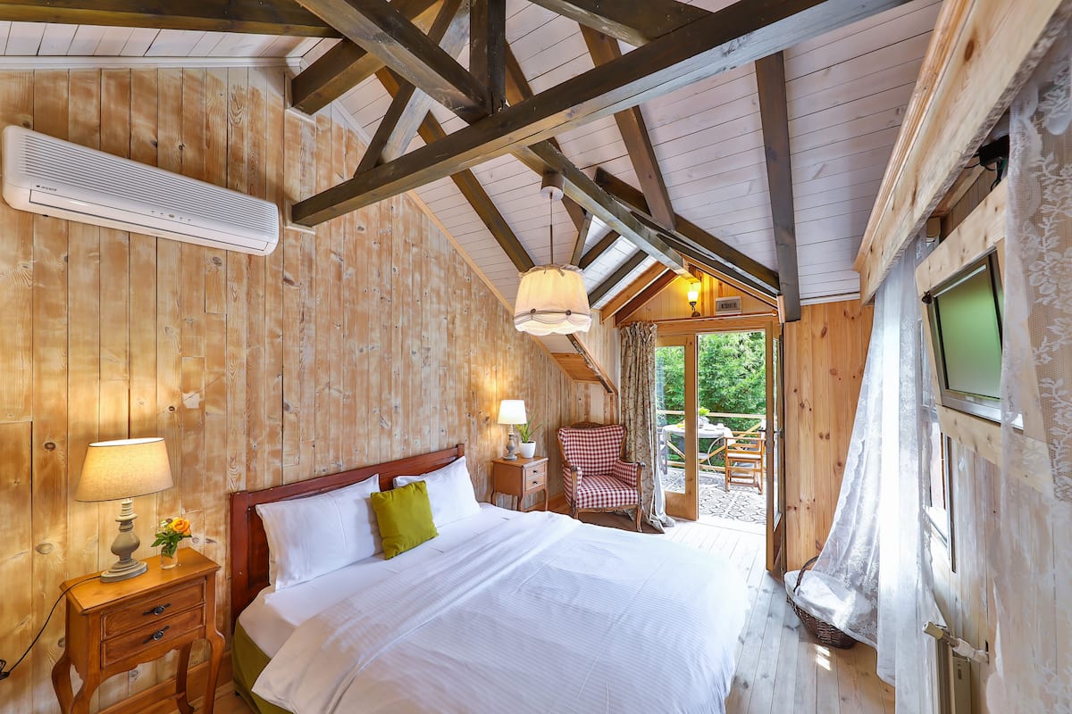 A wooden cabin bedroom with a bed, bedside table, lamp, chair, and a view of the outdoors.