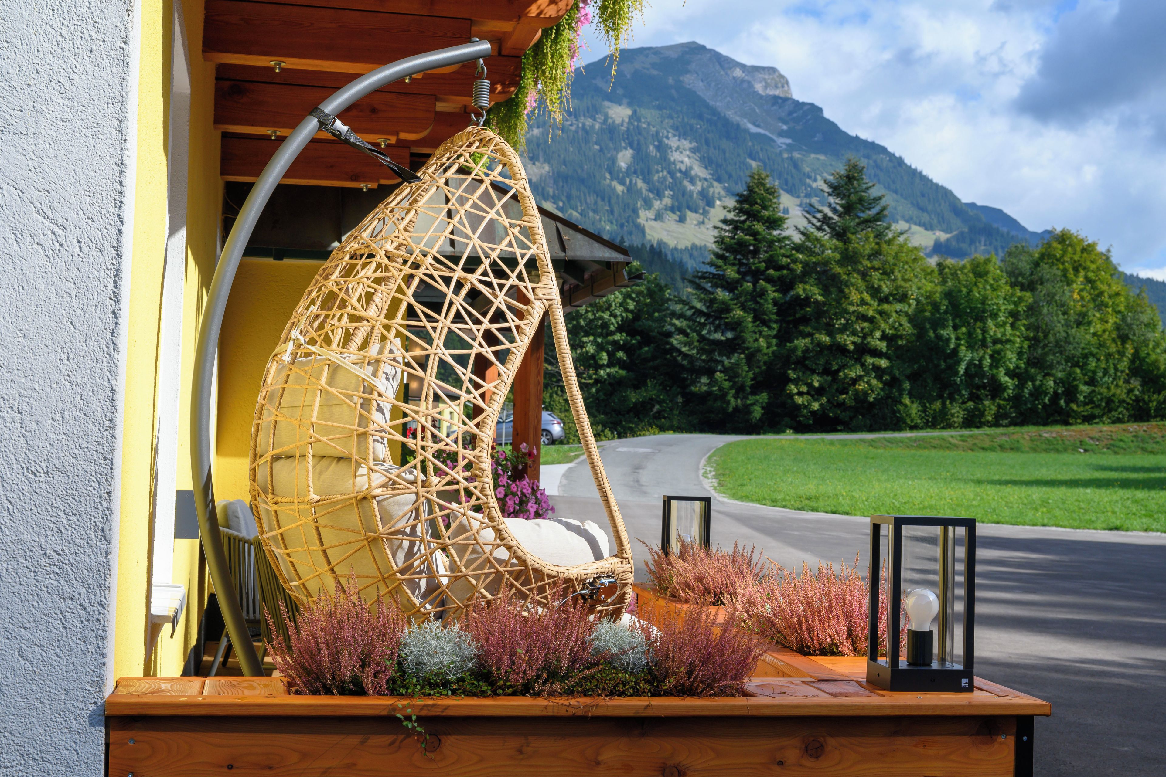 A wicker chair with a cushion, a wooden bench, and a potted plant with pink flowers.