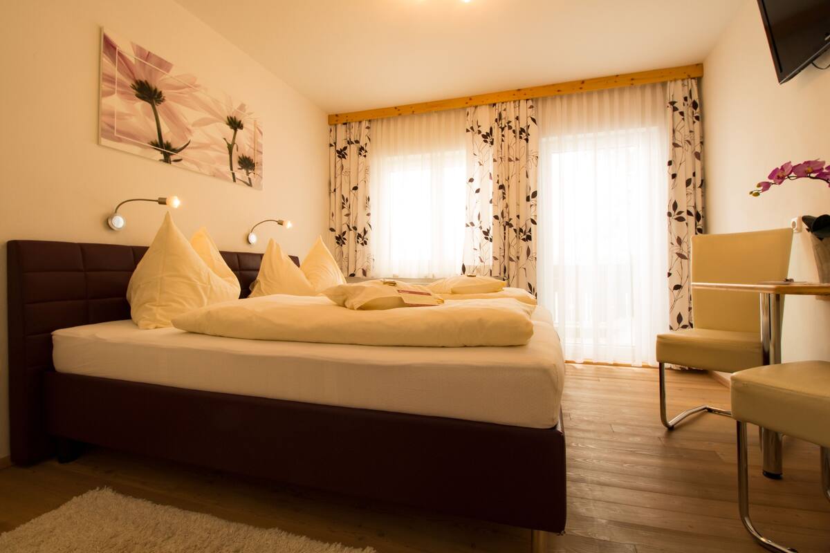 Une chambre à coucher avec un lit, une chaise, un bureau, une télévision et une fenêtre avec des rideaux.