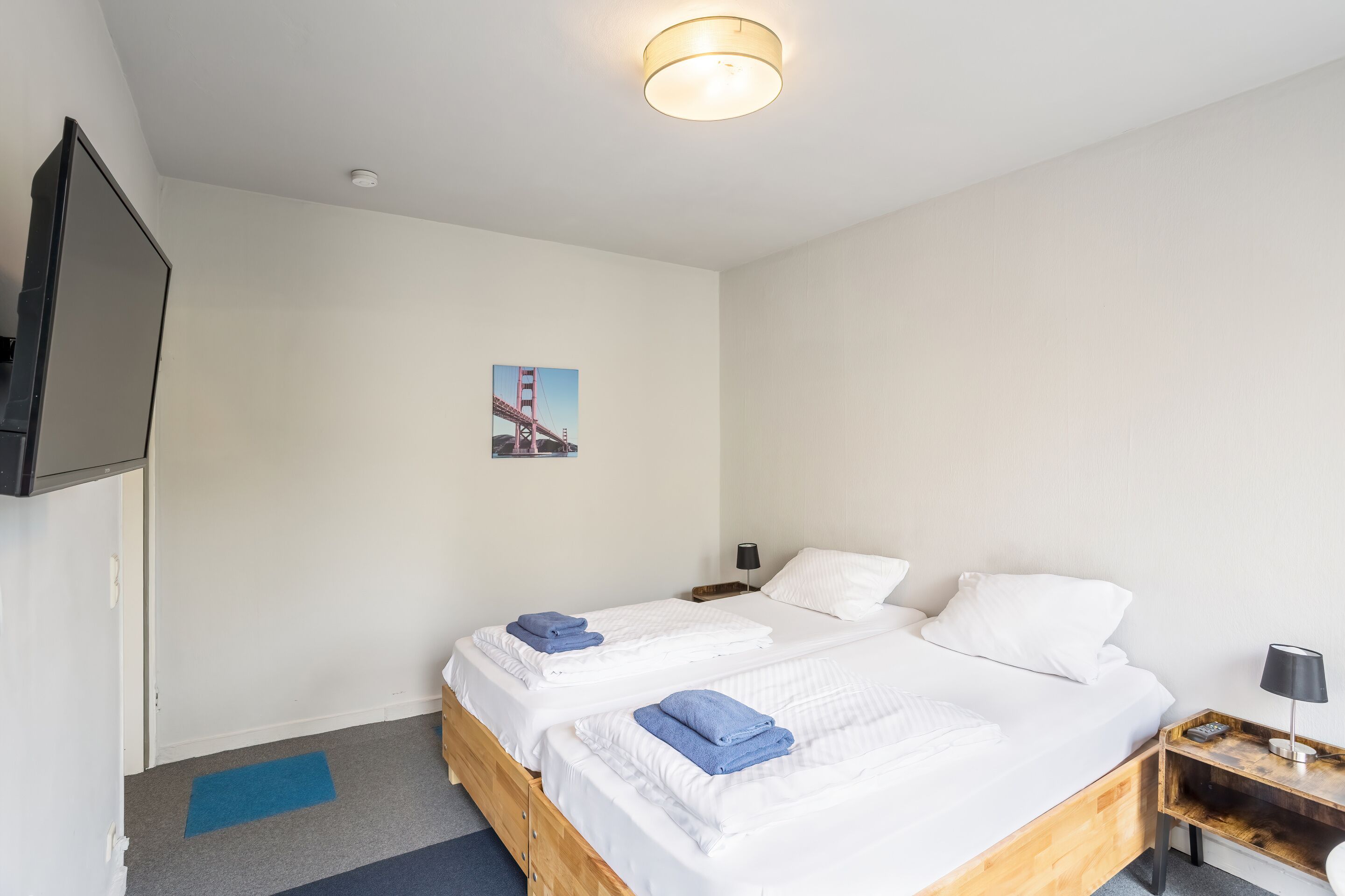 A hotel room with two beds, a flat-screen TV, a nightstand with a lamp, and a picture of the Golden Gate Bridge on the wall.