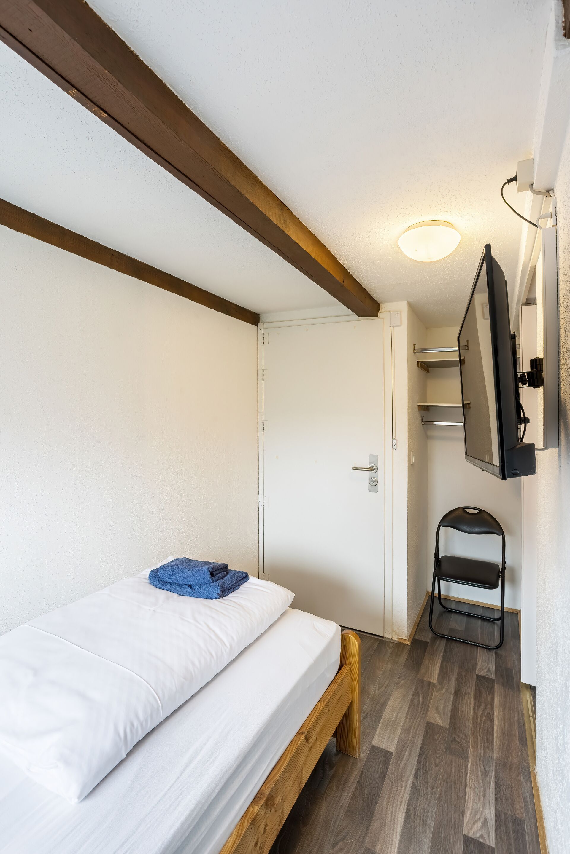 A small, compact room with a bed, a chair, a television mounted on the wall, and a door leading to a bathroom.