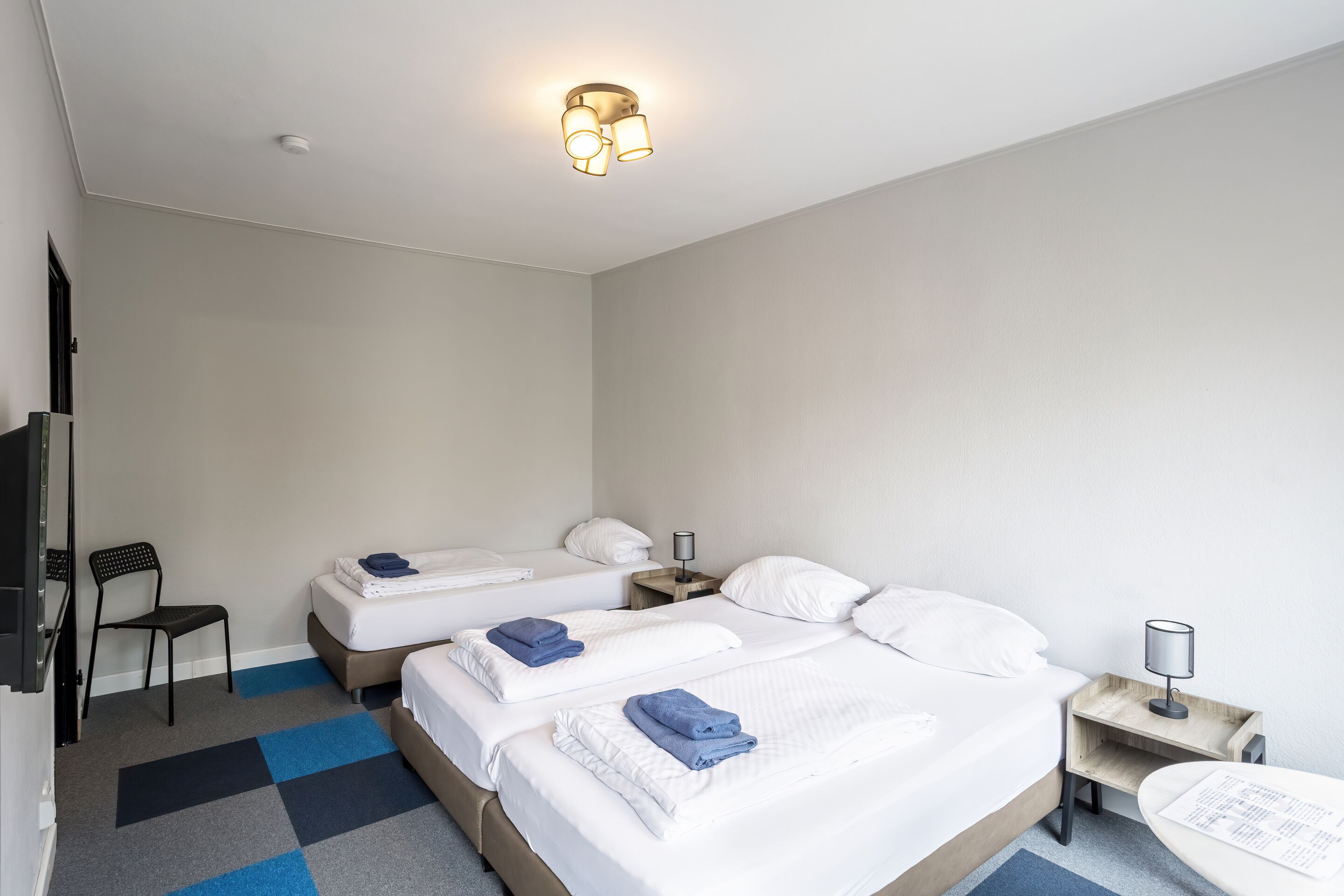 A hotel room with two beds, a desk, a chair, and a small table with a lamp.