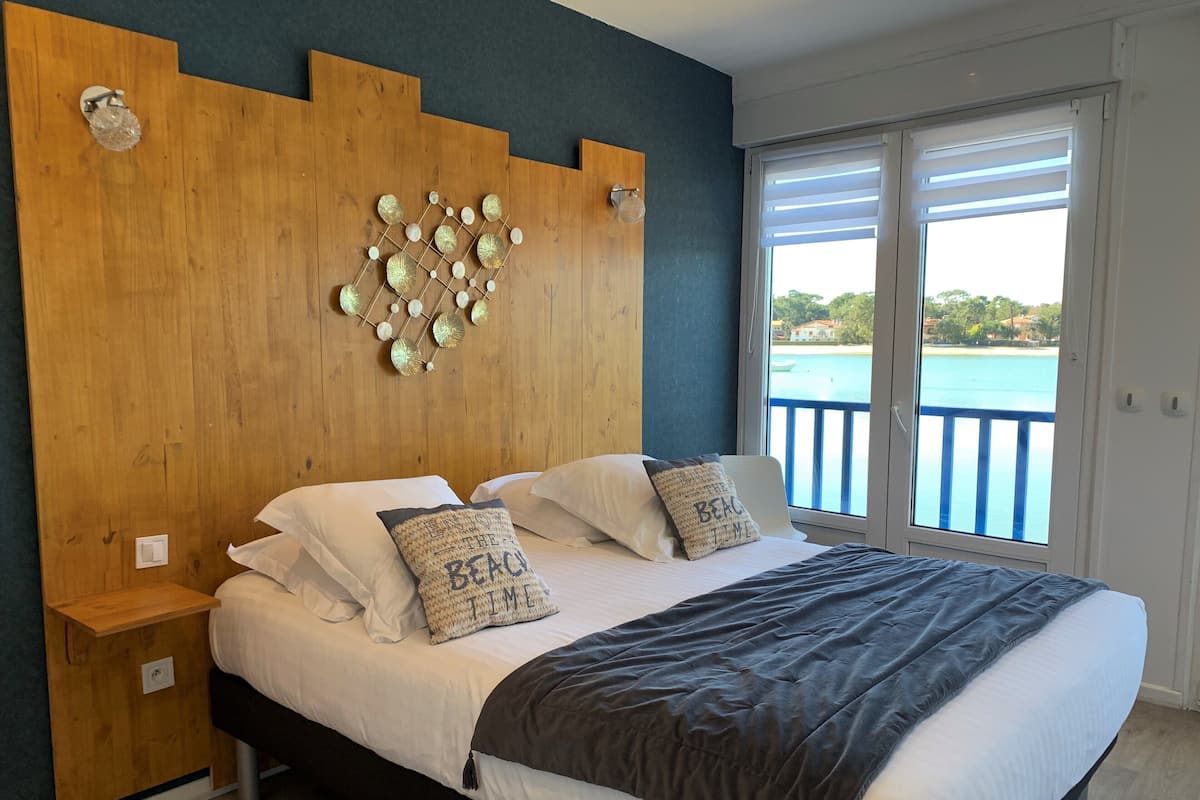 A bedroom with a wooden headboard, a large bed with white and patterned pillows, a dark grey blanket, and a view of a blue body of water through a balcony.