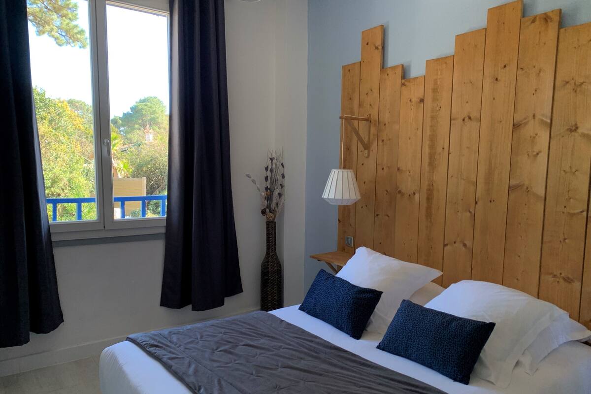 A bedroom with a wooden headboard, a bed with white and dark blue pillows, a bedside lamp, and a view of greenery through a window.