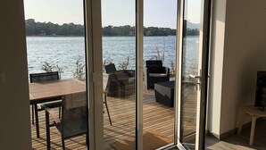 Una habitación con puerta corrediza de vidrio que da a una terraza exterior con vistas a un lago y árboles.
