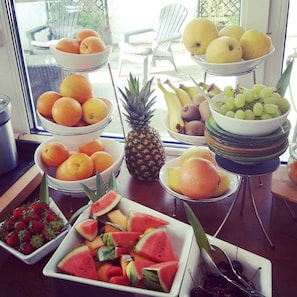 A fruit and vegetable display with various fruits like oranges, grapes, and watermelon, arranged on a table in a room with a view of outdoor seating.
