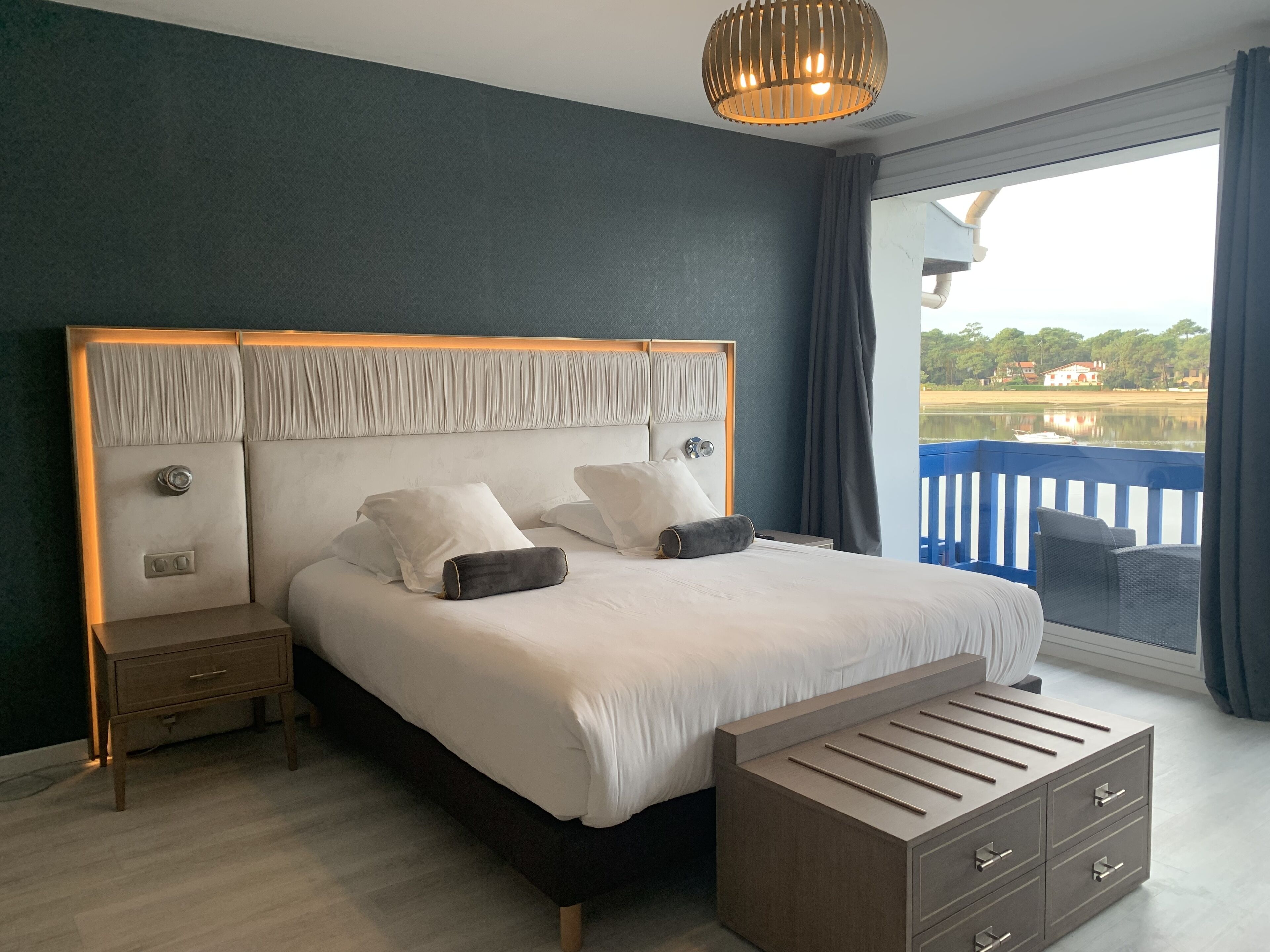 A bedroom with a large bed, a bedside table, a nightstand, a dresser, and a view of the outdoors.