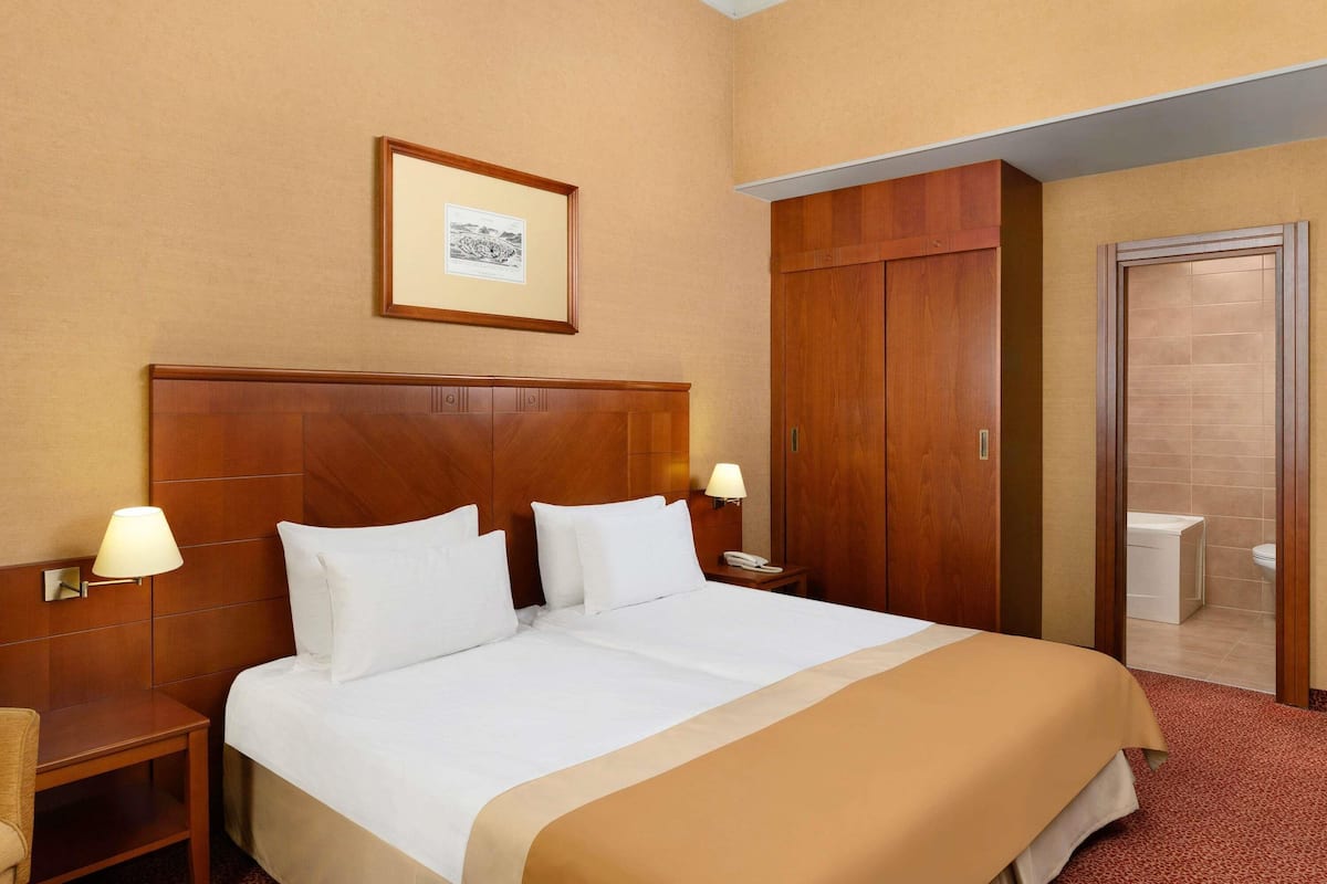 Ein Hotelzimmer mit einem groĂen Bett, zwei Nachttischlampen, einem Holzschrank, einem gerahmten Bild an der Wand und einem durch eine offene TĂŒr einsehbaren Badezimmer.