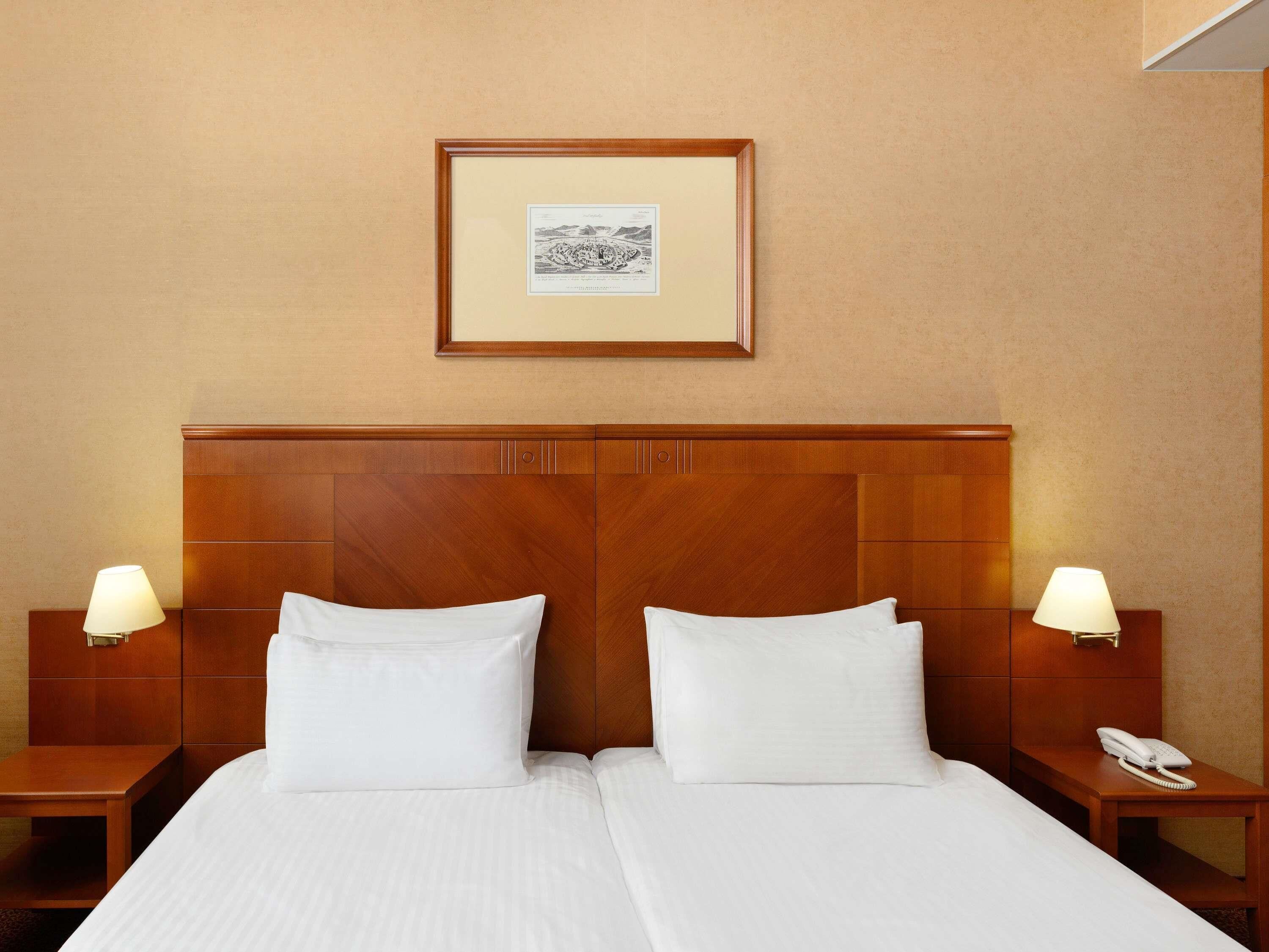 A hotel room with a wooden headboard, two white pillows, a framed picture on the wall, and bedside lamps.