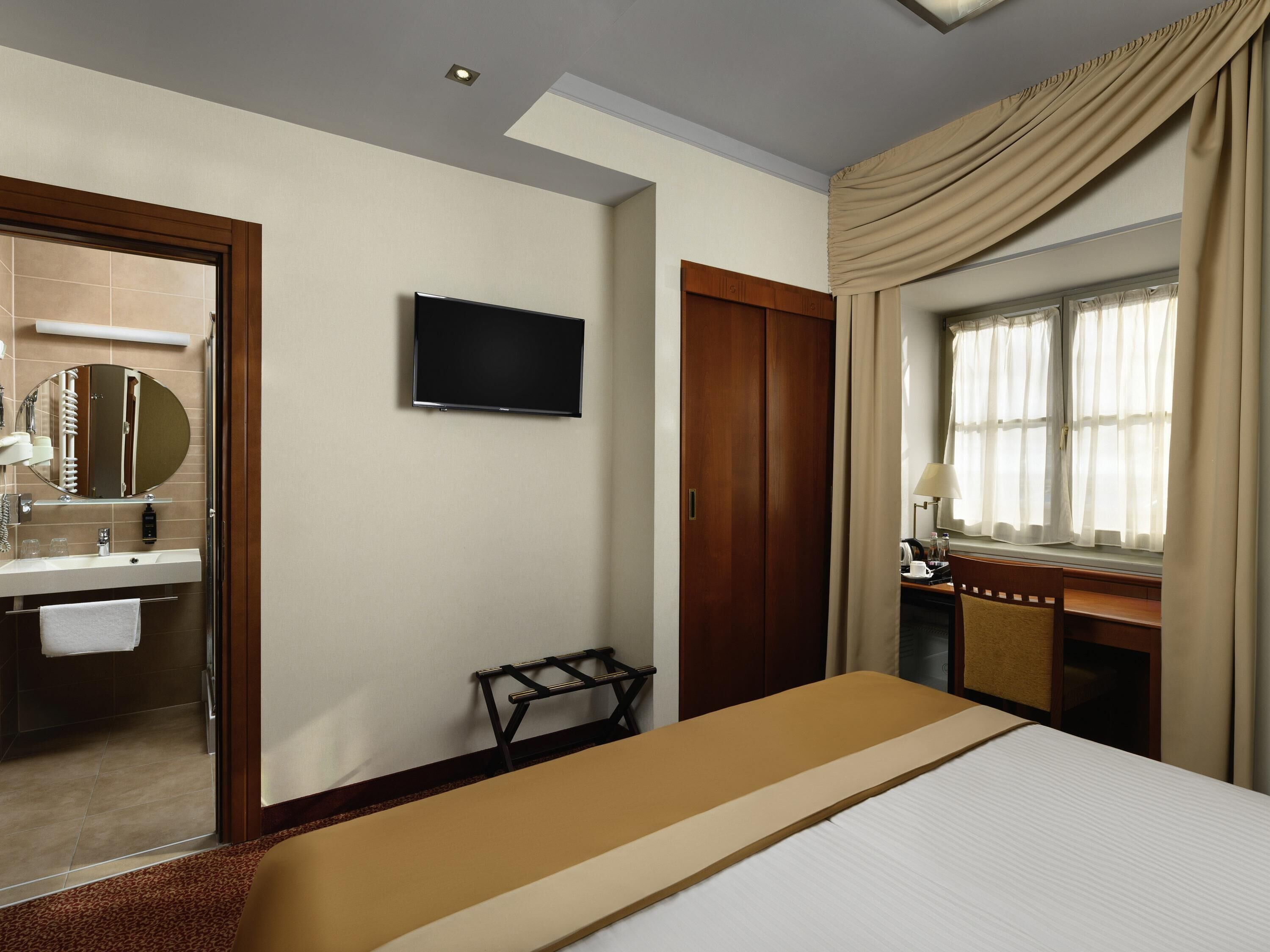 Habitación de hotel con cama, televisión, escritorio, ventana con cortinas, baño con espejo y perchero.