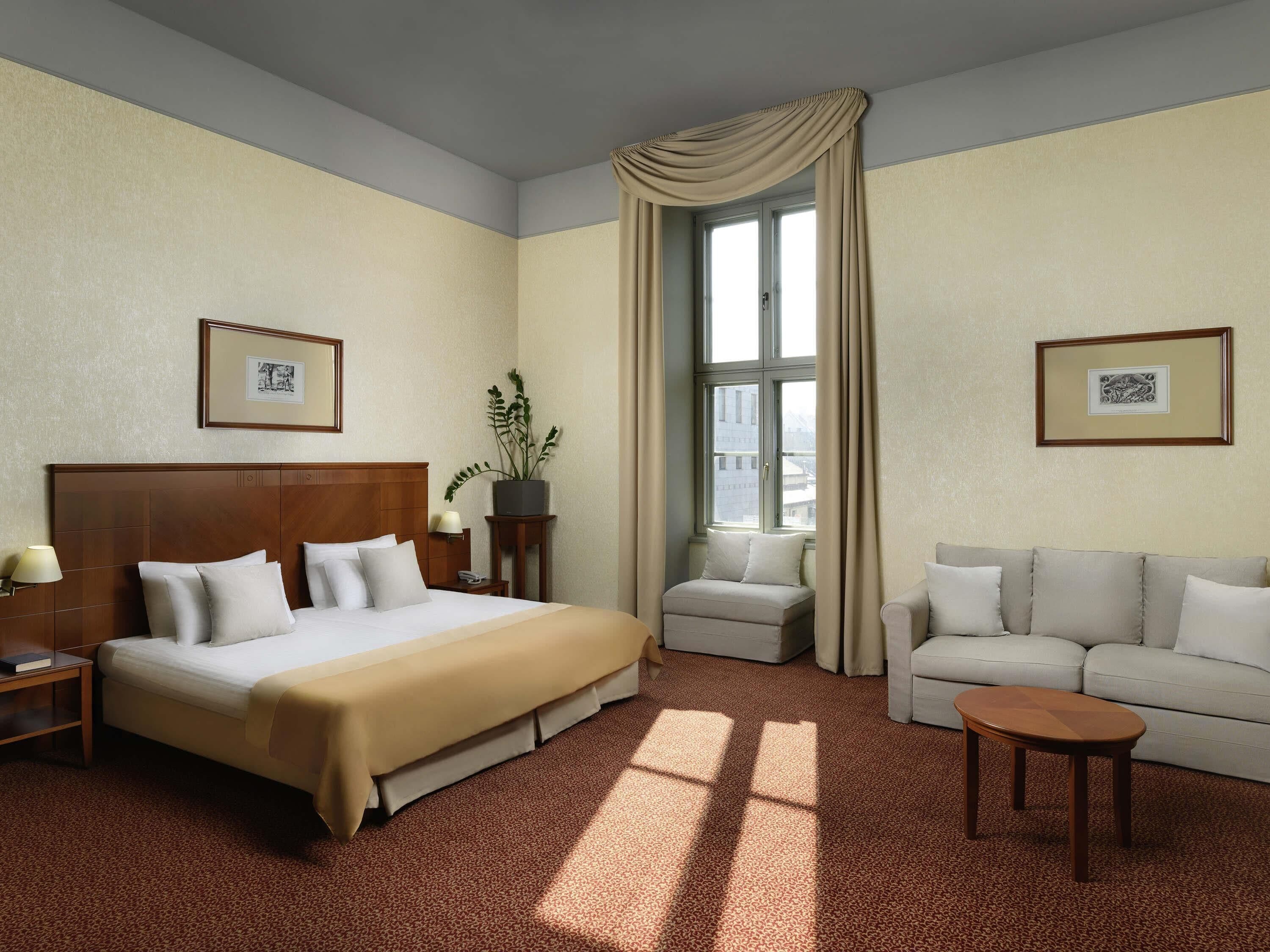 Un cuarto de hotel con una cama grande, un sofá, una mesita, una mesita de noche, una planta en maceta y dos cuadros en las paredes.